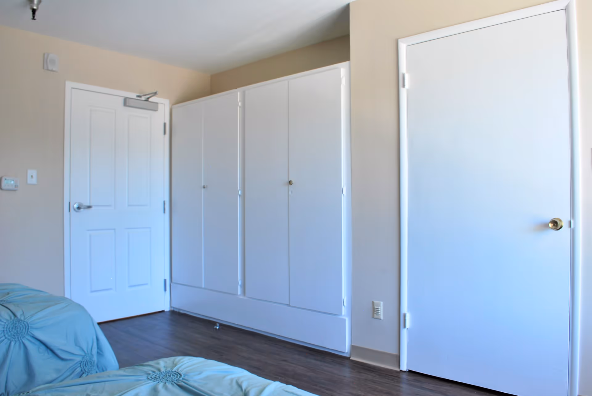 Bedroom interior with two white doors, built-in closets, and blue-covered beds on dark wood flooring.