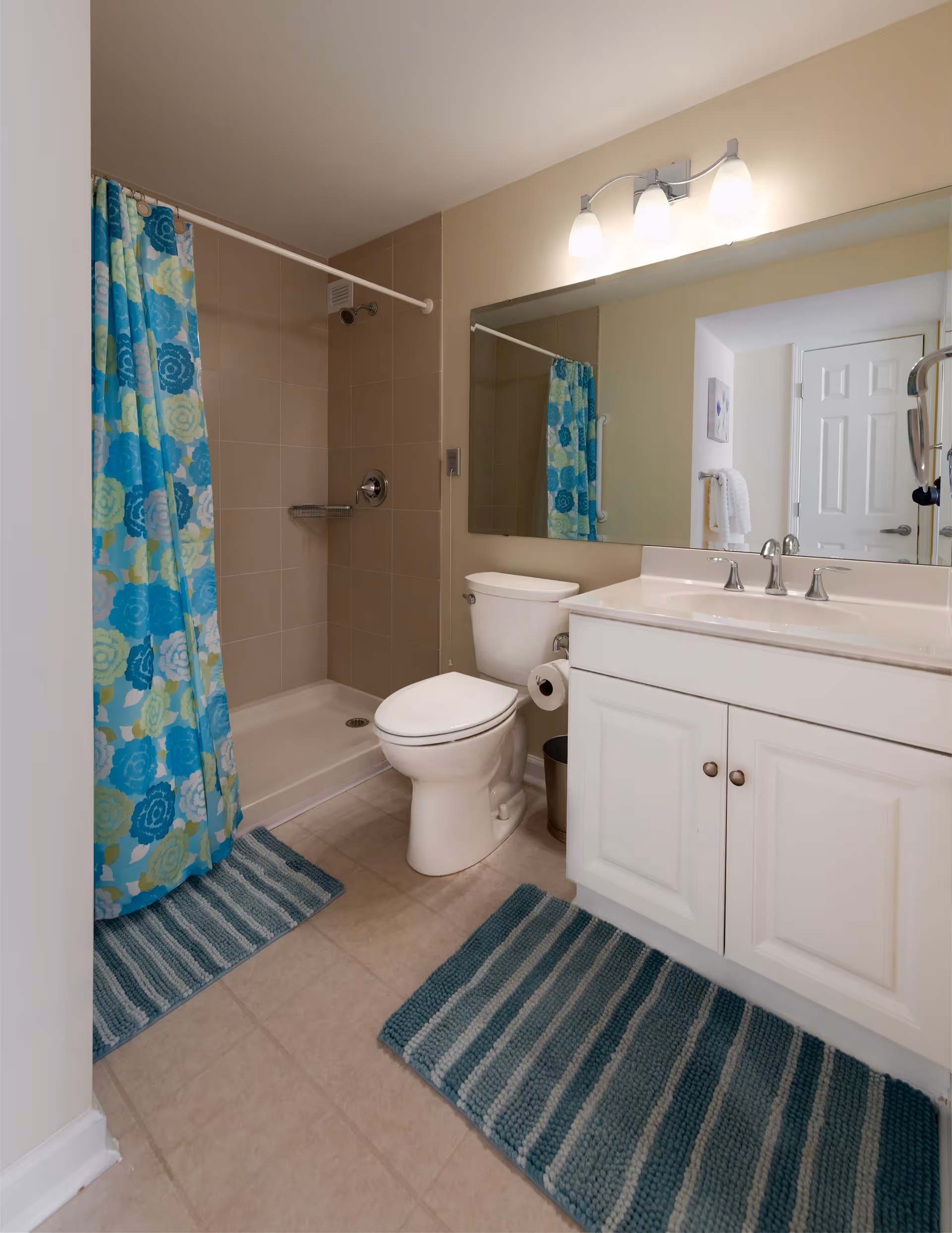 A clean bathroom featuring a white toilet, a white vanity with a sink and silver faucet, a large mirror above the sink, and a shower area with beige tiles and a colorful blue and green floral shower curtain. Two blue striped bath mats are placed on the tiled floor.