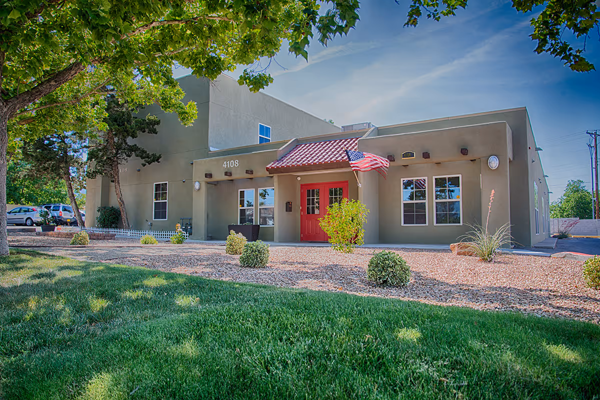 Exterior view of a single-story building with a red door, beige walls, and an American flag mounted near the entrance. The building is surrounded by a landscaped area with green grass, small bushes, and trees under a clear blue sky.