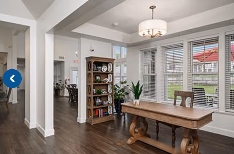 A bright interior room with large windows featuring white blinds, a wooden table with a plant on top, a wooden chair, and a tall wooden bookshelf with decorative items and books. The room has wooden flooring and a modern ceiling light fixture.
