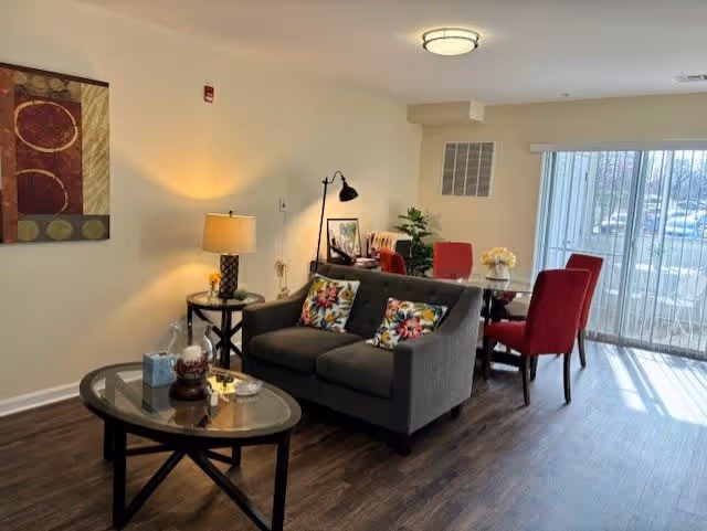 A cozy living and dining area with a gray sofa adorned with floral pillows, a glass coffee table, a side table with a lamp, and a dining table with red chairs near a large window with vertical blinds letting in natural light.