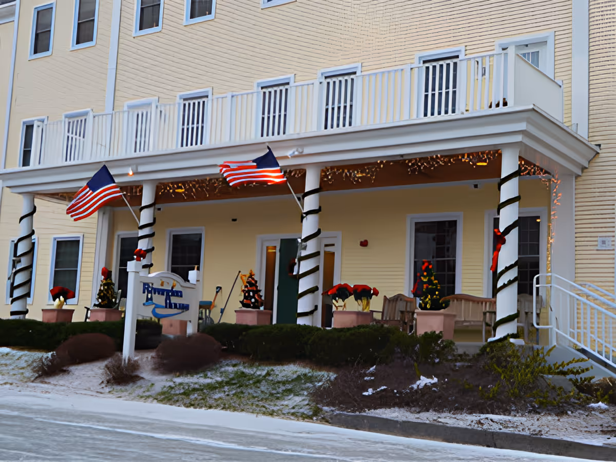 Front exterior view of a yellow senior living facility building with a porch decorated with American flags, Christmas trees, and poinsettia plants. The porch has white railings and columns wrapped with garlands, and there is a sign in front of the building.