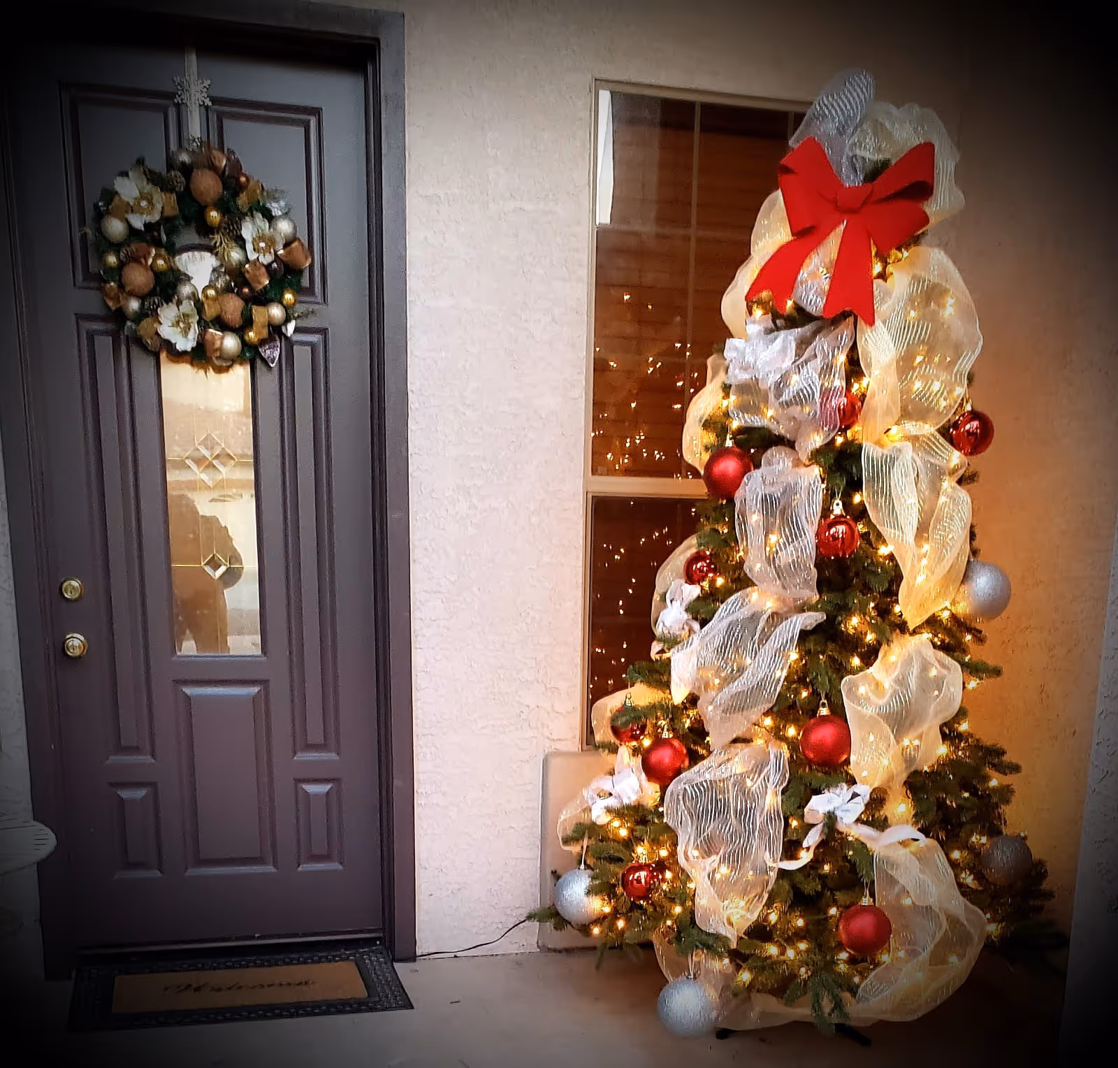A decorated Christmas tree with white mesh ribbon, red and silver ornaments, and lights stands next to a dark brown front door with a festive wreath. The door has a glass panel with a decorative design, and there is a welcome mat on the floor.