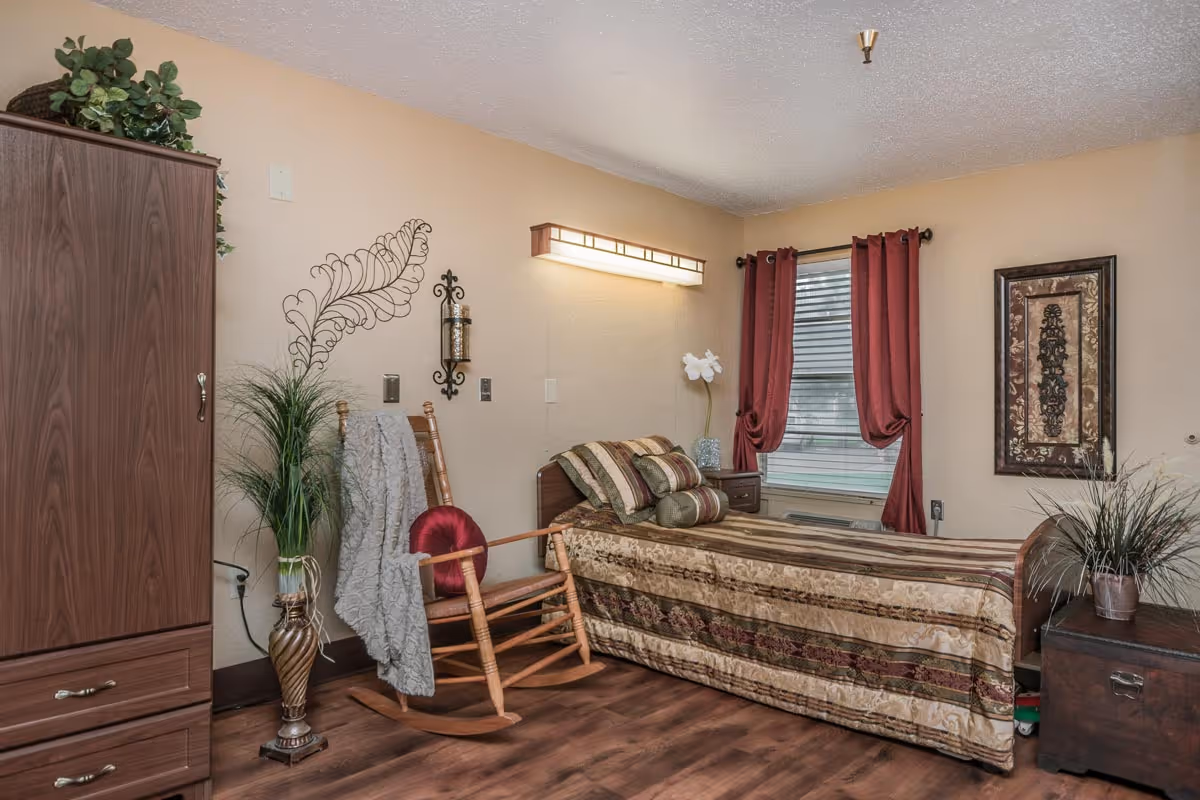 A cozy single bedroom featuring a twin bed with a patterned bedspread, a wooden rocking chair, armoire, decorative plants, and a window with red curtains.