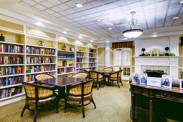 Bright library-style common room with built-in bookshelves, tables and upholstered chairs near a fireplace.