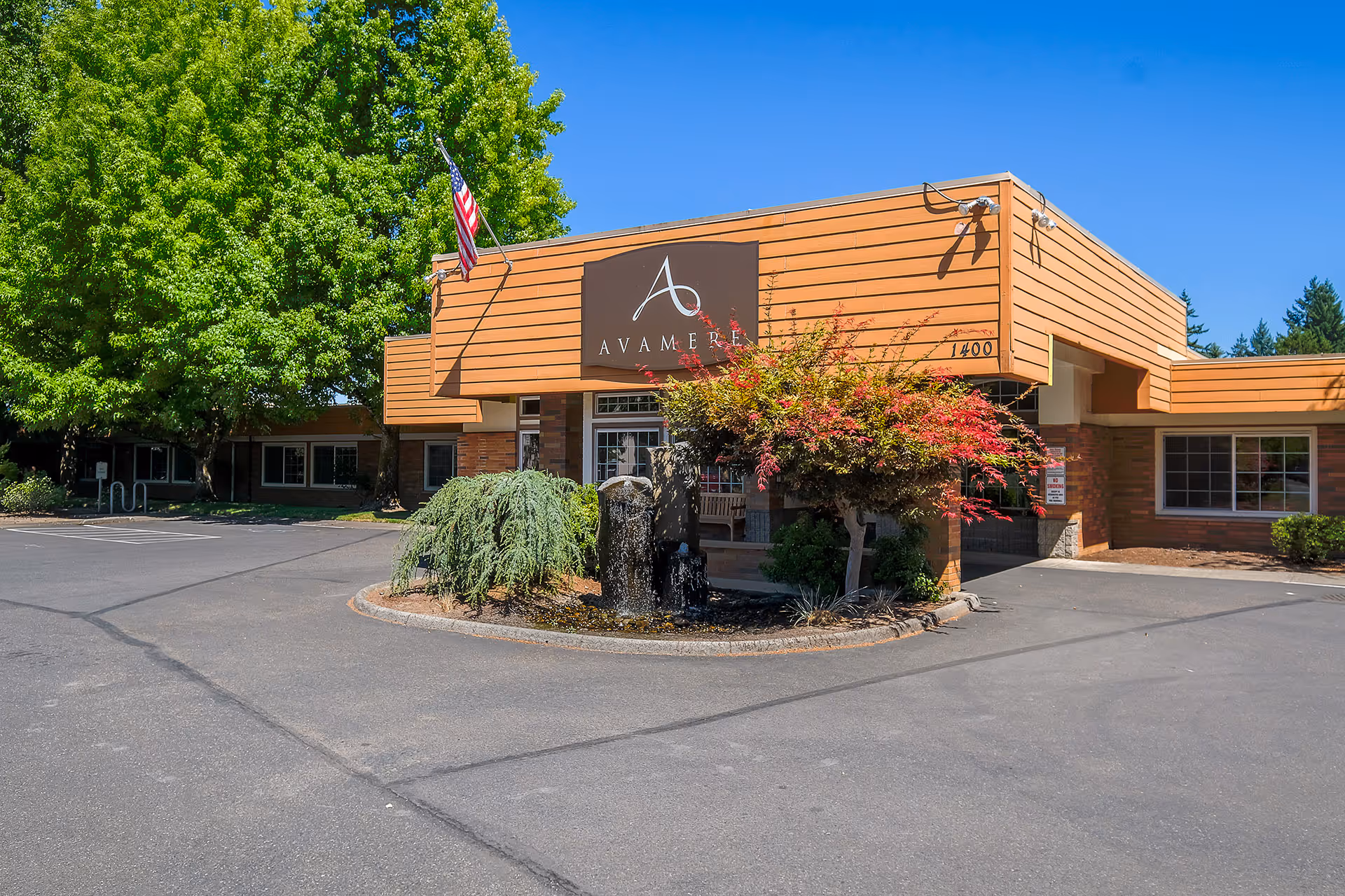 Exterior view of Avamere Rehabilitation of Oregon City building with a brown and orange facade, an American flag, and landscaping including trees and shrubs under a clear blue sky.