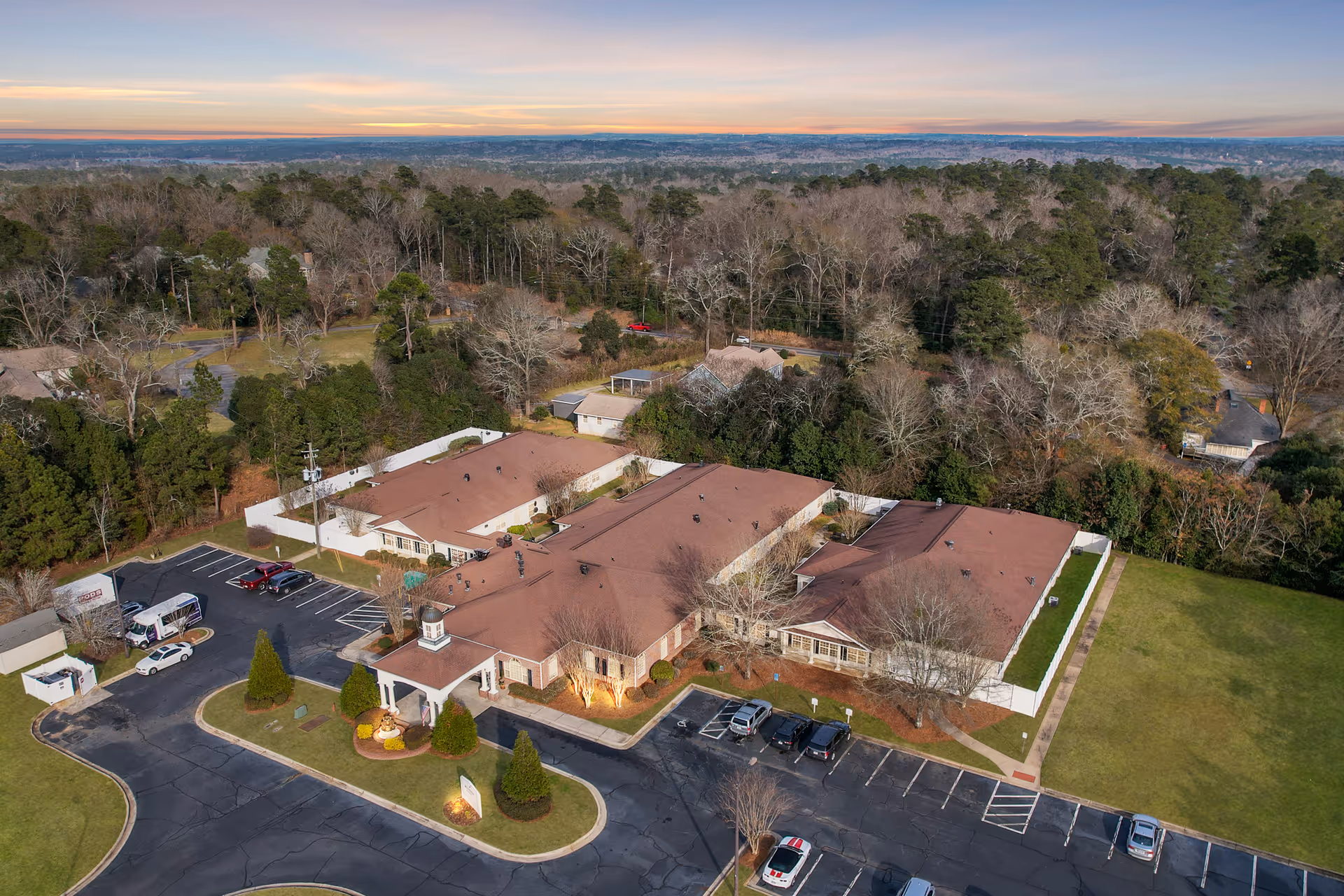 Aerial view of PruittPlace - Wesleyan senior living facility surrounded by trees and greenery, with a parking lot and several cars in front of the building under a clear sky at sunset.