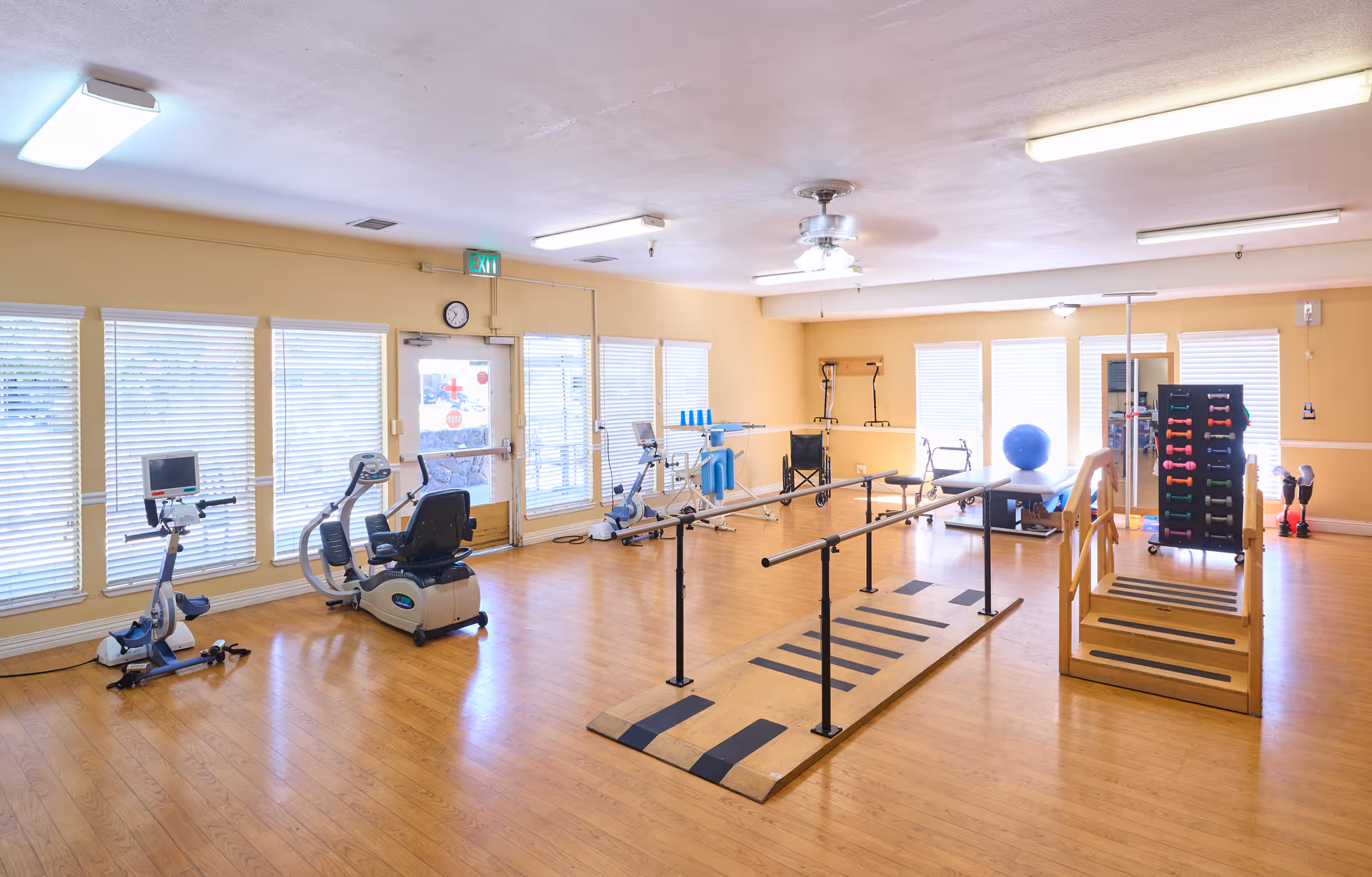 A spacious rehabilitation or physical therapy room with exercise equipment including stationary bikes, parallel bars for walking exercises, a set of stairs for step training, a rack of colorful dumbbells, a large blue exercise ball, and a wheelchair. The room has wooden flooring, large windows with blinds, and bright overhead lighting.