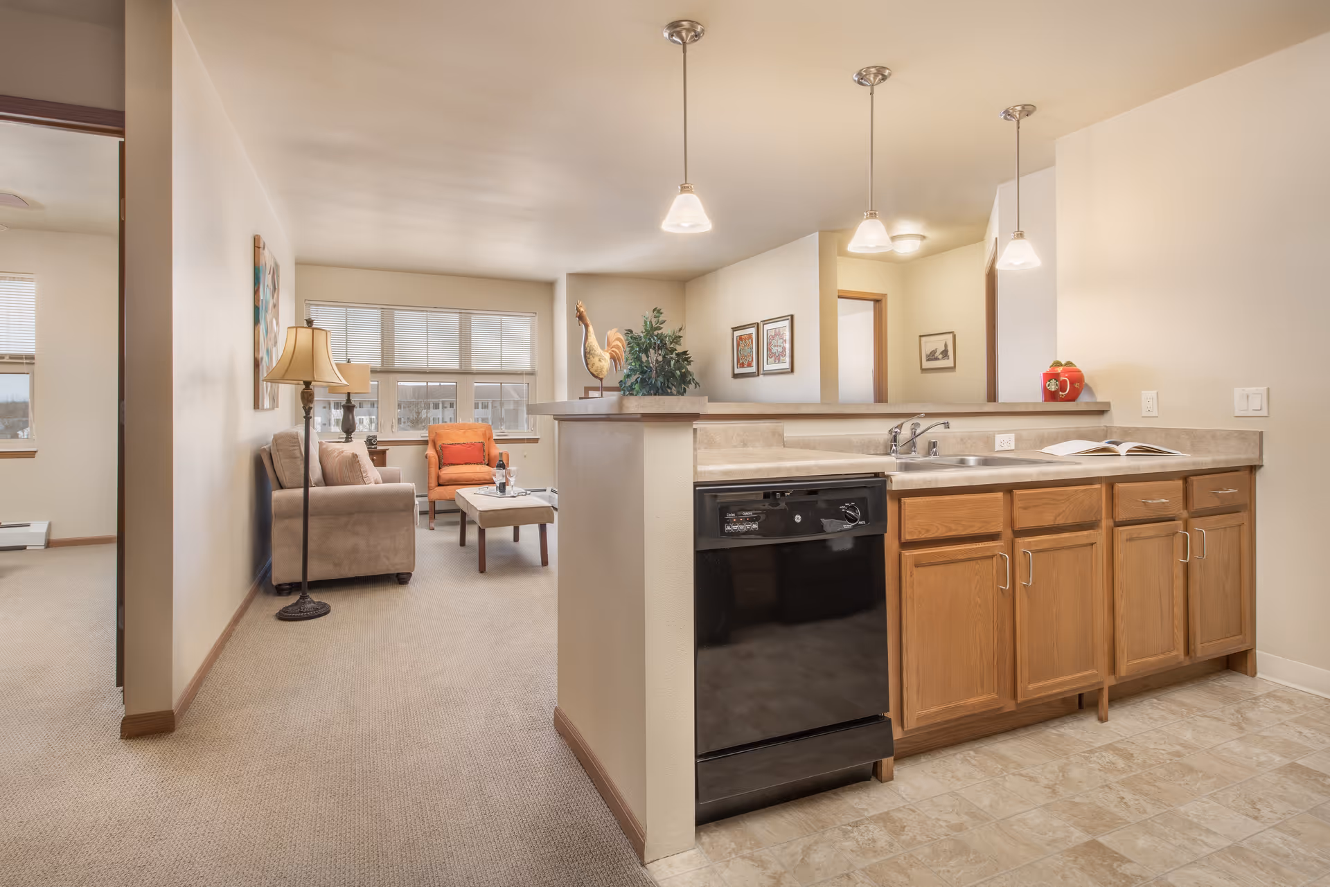 Interior view of a senior living facility apartment showing a kitchen area with wooden cabinets, a black dishwasher, and a countertop with a sink. The kitchen opens to a living room furnished with a beige sofa, an orange armchair, a coffee table, and a floor lamp. Large windows in the living room allow natural light to fill the space.