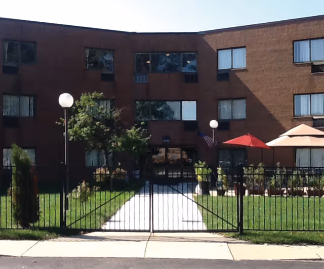 Front exterior view of a three-story brick building with multiple windows, a gated entrance, a concrete walkway leading to the main door, green lawn, trees, and patio umbrellas on the right side.