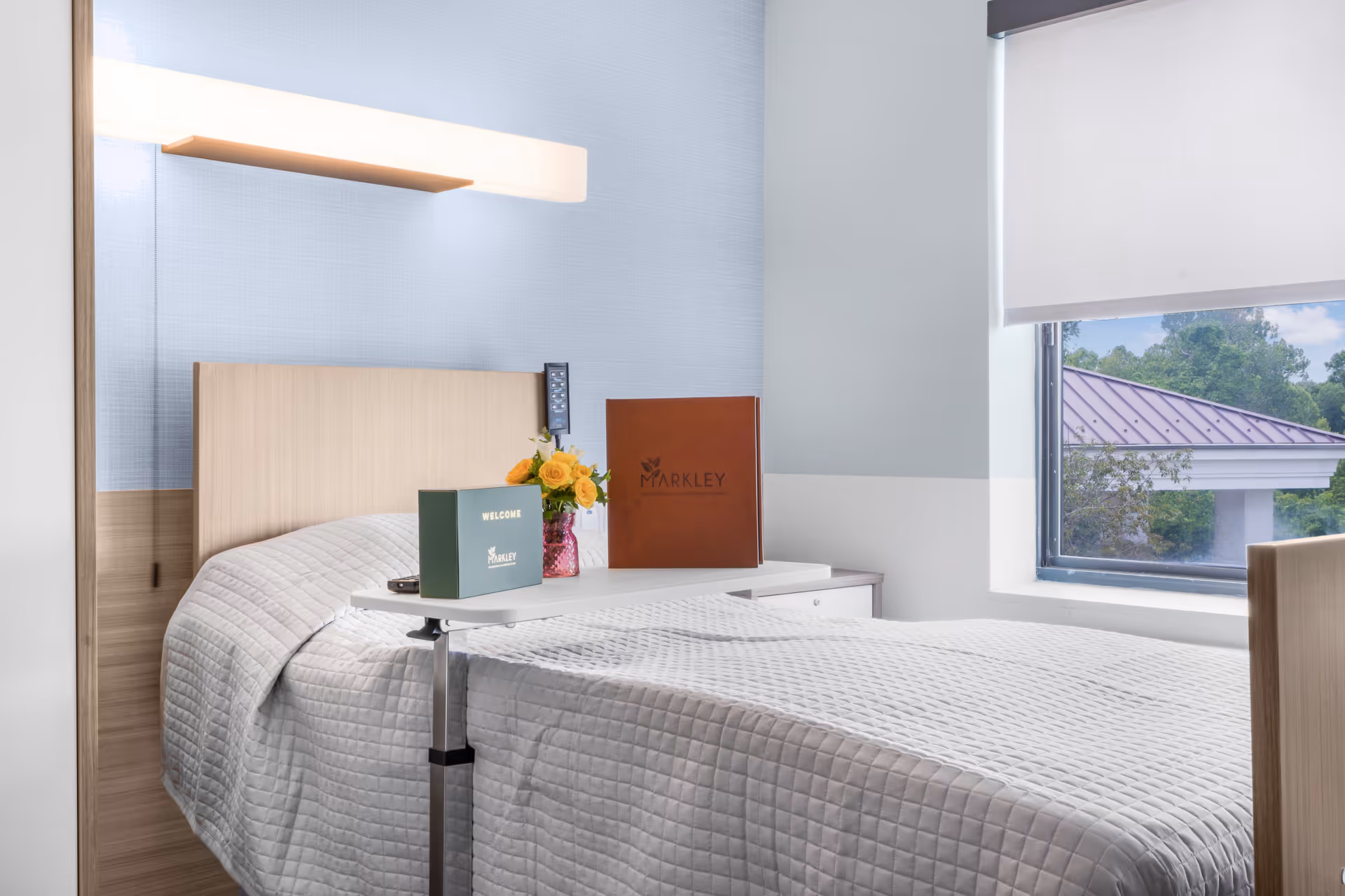 A neatly made single bed in a healthcare facility room with a light wooden headboard and a white quilted bedspread. A small overbed table holds a welcome card, a brown folder with the Markley logo, and a vase with yellow flowers. A large window with a white roller shade partially open shows greenery and a building roof outside.