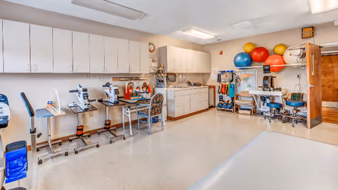 A bright therapy or rehabilitation room with exercise equipment, therapy balls, chairs, cabinets, and a map on the wall. The room has a clean, organized appearance with white walls and ceiling lights.