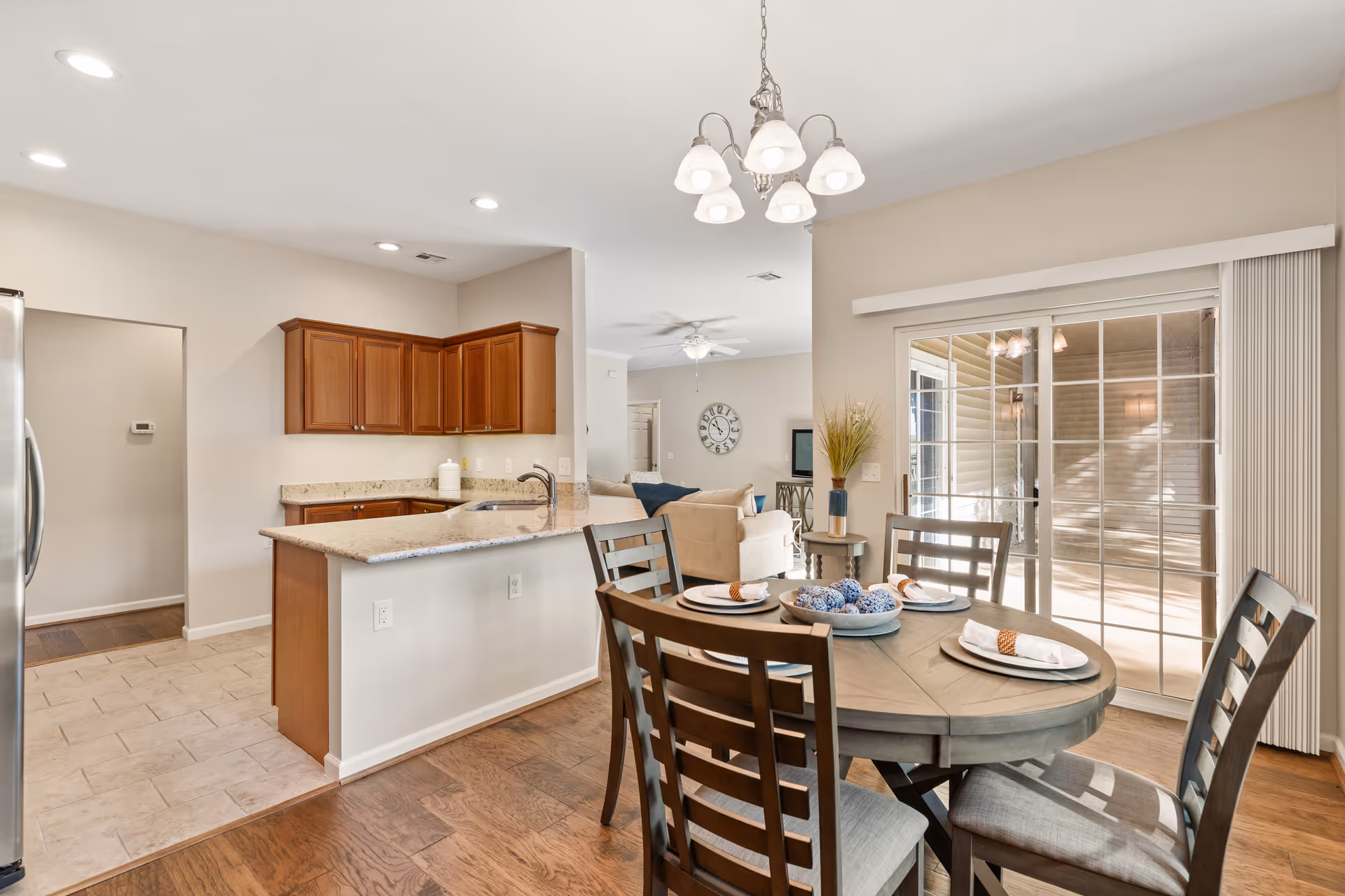 A bright and spacious dining area with a round wooden table set for four, adjacent to a kitchen with granite countertops and wooden cabinets. Sliding glass doors lead to an outdoor patio, and a living room with a beige sofa and wall clock is visible in the background.