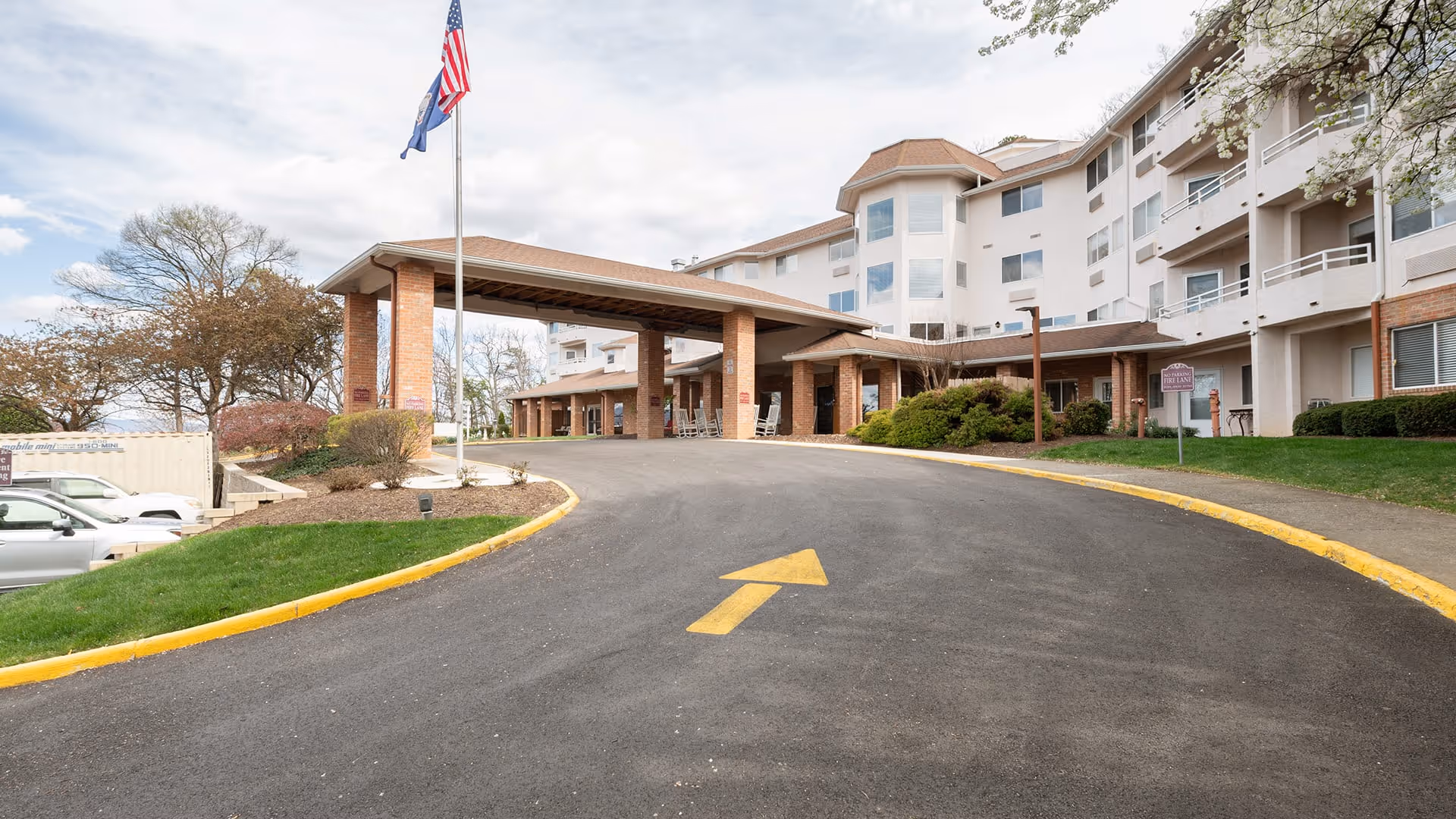 Front exterior view of Holiday Elm Park Estates, showing a multi-story building with balconies, a covered entrance with brick pillars, an American flag and another flag on flagpoles, a curved driveway with a yellow arrow painted on the asphalt, and some parked cars and landscaping with trees and bushes.