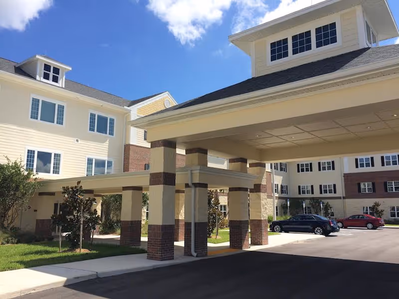Exterior view of a senior living facility showing a covered entrance with multiple pillars and a driveway. The building has multiple windows and a combination of light yellow siding and brick accents. There are a few cars parked in the parking lot under a partly cloudy blue sky.