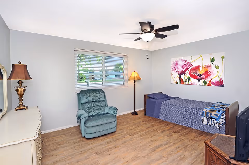 A simple bedroom in an assisted living facility featuring a single bed with blue bedding and a colorful floral painting above it. There is a green recliner chair near a window with blinds, two lamps providing warm light, a ceiling fan, and wooden flooring.