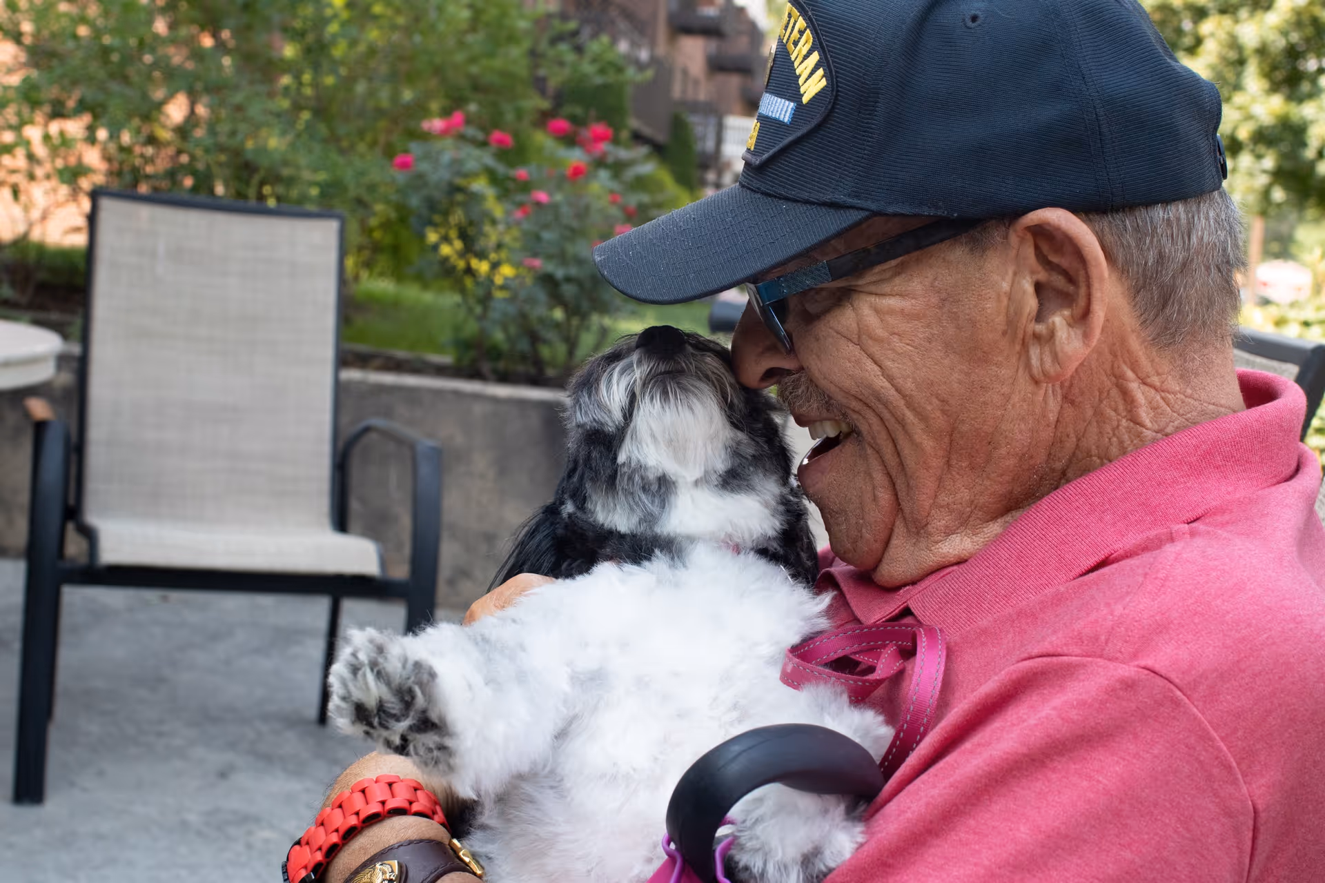An elderly man wearing a black veteran cap and pink shirt is smiling and holding a small black and white dog close to his face outdoors, with garden plants and a chair in the background.
