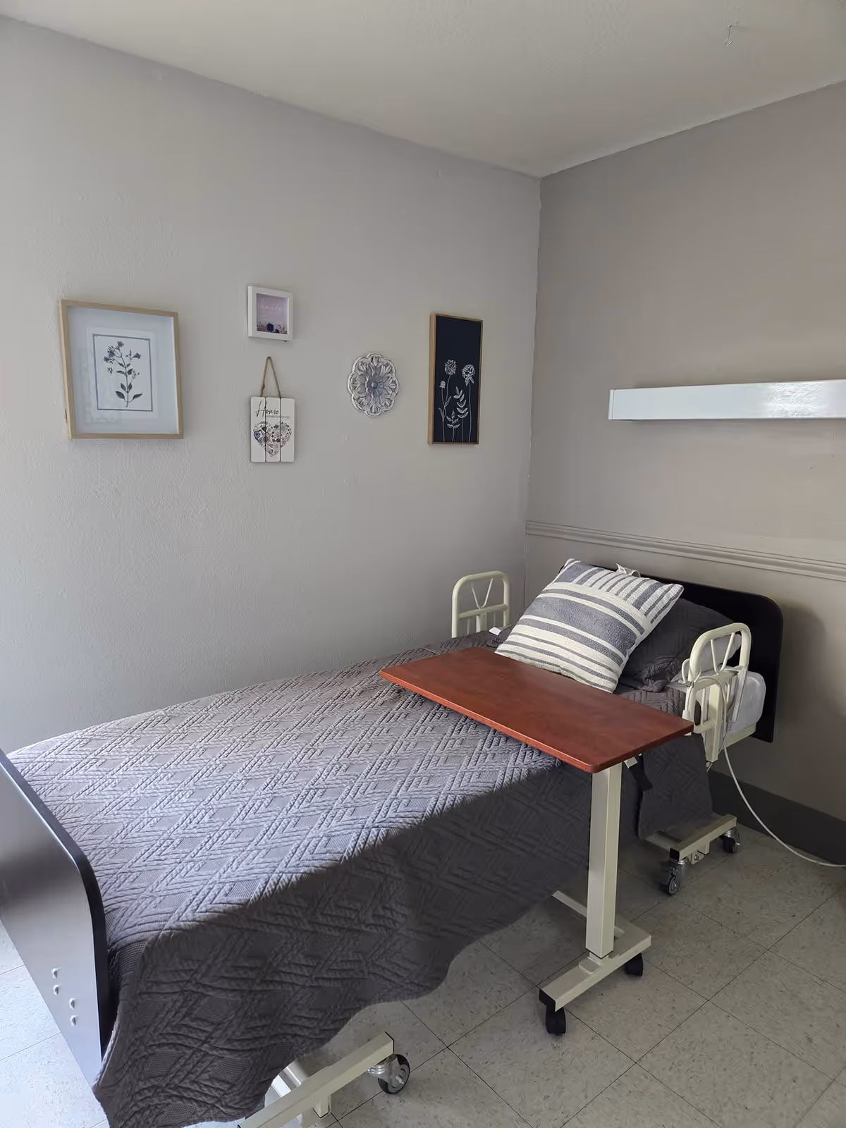 A simple room with a single hospital-style bed covered with a gray quilted blanket and a striped pillow. A wooden overbed table is positioned over the bed. The walls are light gray and decorated with four small framed pictures and wall hangings.