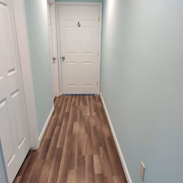 A narrow hallway with light blue walls and wood-patterned flooring leading to a white door labeled with the number 6. There are additional white doors on the left side of the hallway.