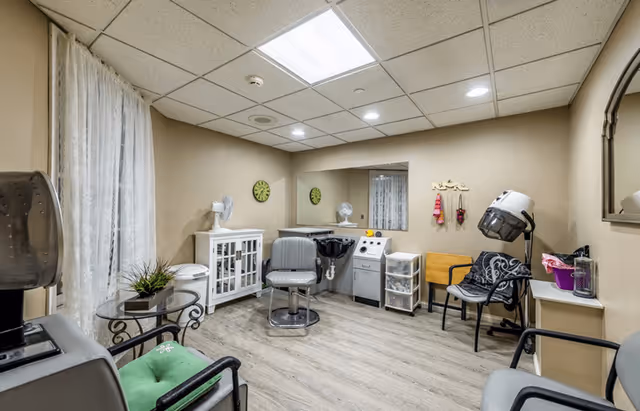 A small hair salon room in an assisted living facility with a salon chair in front of a wash basin, a hair dryer chair, a glass table with a green cushion chair, a white cabinet, a wall mirror, and a clock on the wall. The room has beige walls, wood flooring, and a drop ceiling with fluorescent lighting.