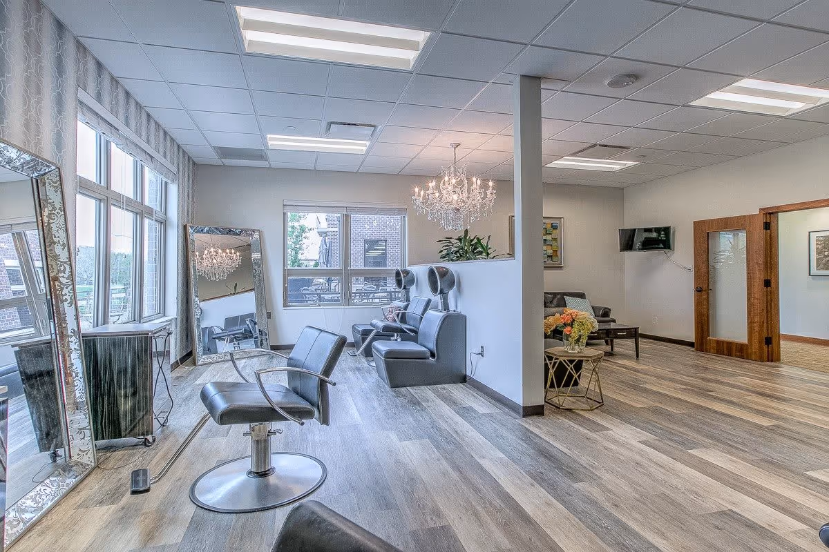 Interior view of a senior living facility's salon area with styling chairs facing large decorative mirrors on the left side. In the background, there are hair drying chairs with hooded dryers, a chandelier hanging from the ceiling, a small seating area with a couch and a coffee table with flowers, and large windows letting in natural light.