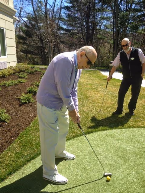 An elderly man wearing sunglasses, a light purple sweater, and white pants is putting a golf ball on a small artificial putting green outside. Another man, also wearing sunglasses and a black vest over a light shirt, stands nearby watching. The scene is set outdoors with grass, trees, and part of a building visible.
