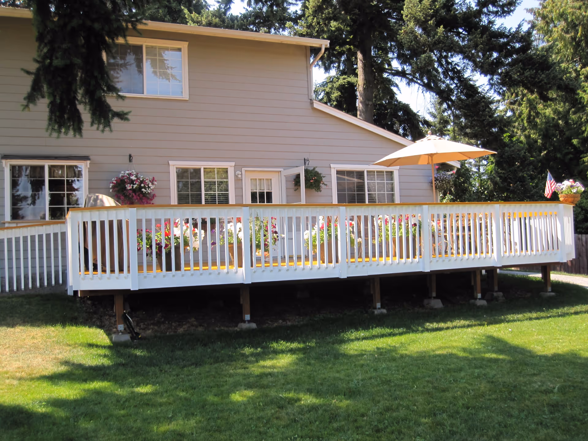 Exterior view of a beige two-story house with a large wooden deck featuring white railings. The deck is decorated with flower pots and has a beige patio umbrella. The house is surrounded by green grass and tall trees.