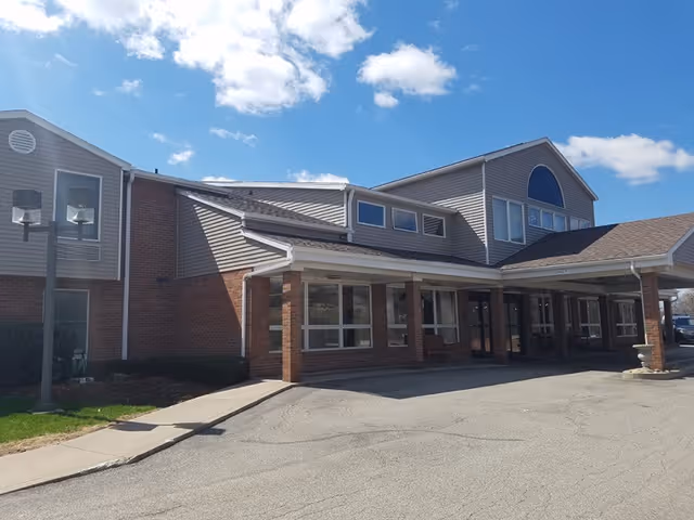 Exterior view of a two-story senior living facility building with brick and siding facade under a blue sky with some clouds. The entrance has a covered drop-off area supported by brick columns and large windows along the front.