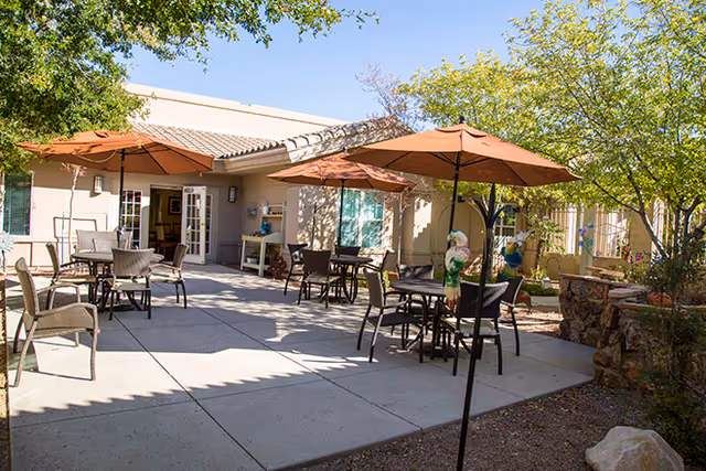 Outdoor patio area at Brookdale Oro Valley with several tables and chairs under large brown umbrellas, surrounded by trees and greenery, adjacent to a building with open double doors.