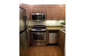 Compact kitchen area with wooden cabinets, stainless steel refrigerator, microwave, oven, and dishwasher. The countertop has a light color and there is a small plant near the sink.