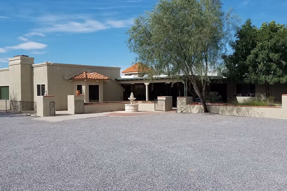 Exterior view of a single-story building with beige stucco walls and a red tile roof section. There is a small courtyard area with a white fountain in the center, surrounded by low walls and some trees providing shade. The sky is clear with a few clouds.