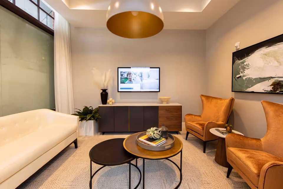 Modern seating area with a white sofa, two orange armchairs, nested coffee tables, a wall-mounted TV, and a cabinet under a pendant light.