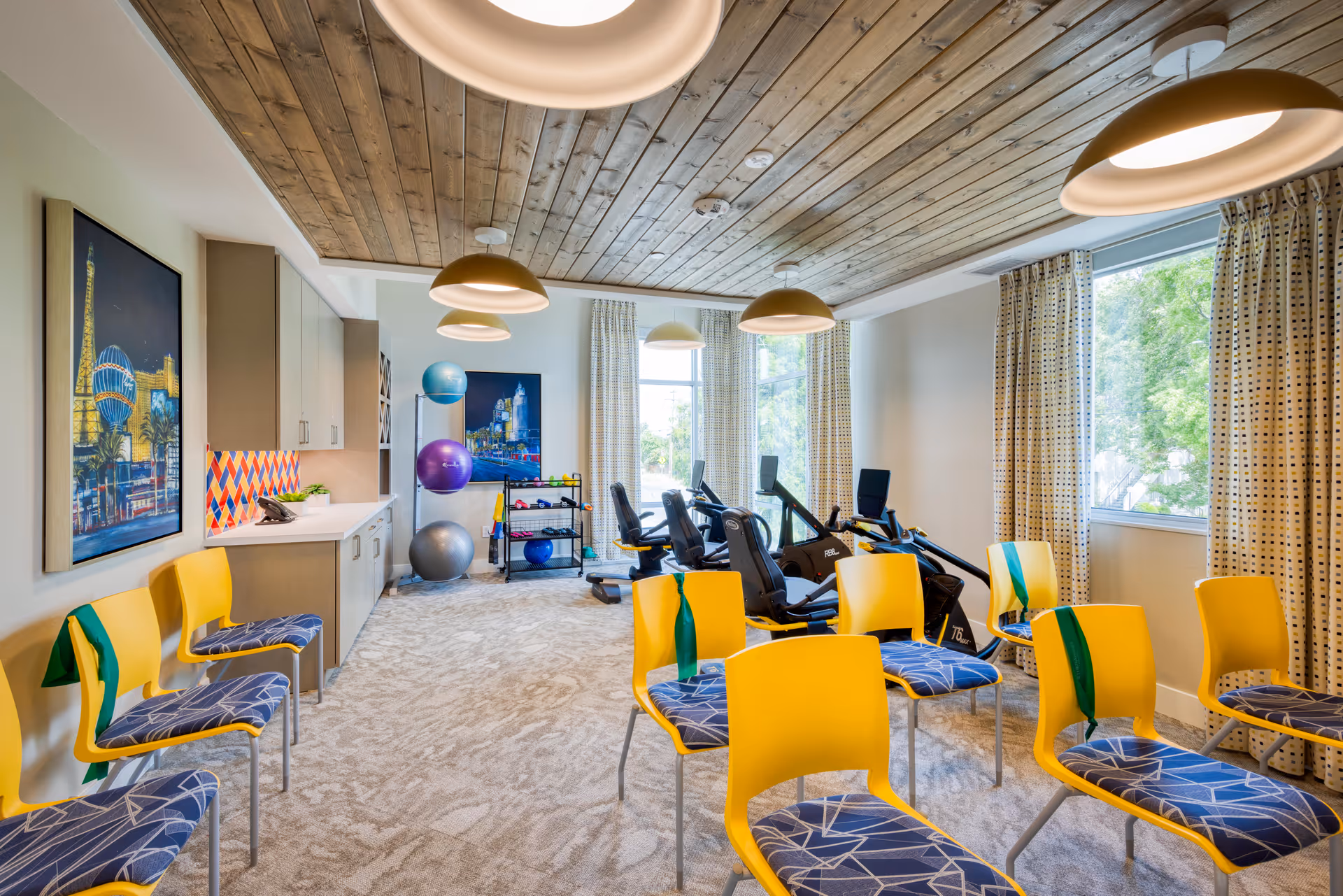 Bright exercise and activity room with yellow chairs, cardio equipment, stability balls, and large windows.