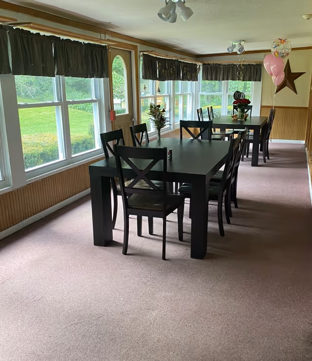 Sunlit dining room with two long dark wooden tables and chairs beside large windows decorated with balloons and an '80' celebration display.