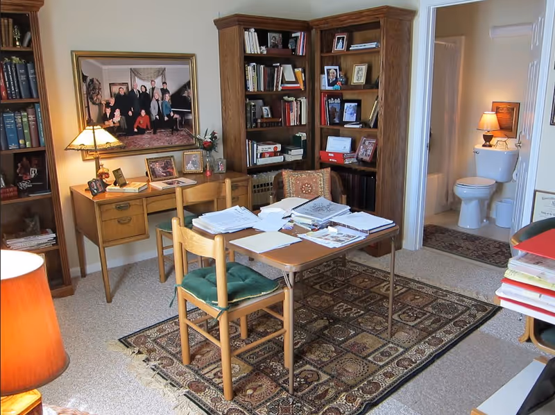 Home office-style living room with a table and chairs covered in papers, bookshelves, and an open door revealing a bathroom with a toilet.