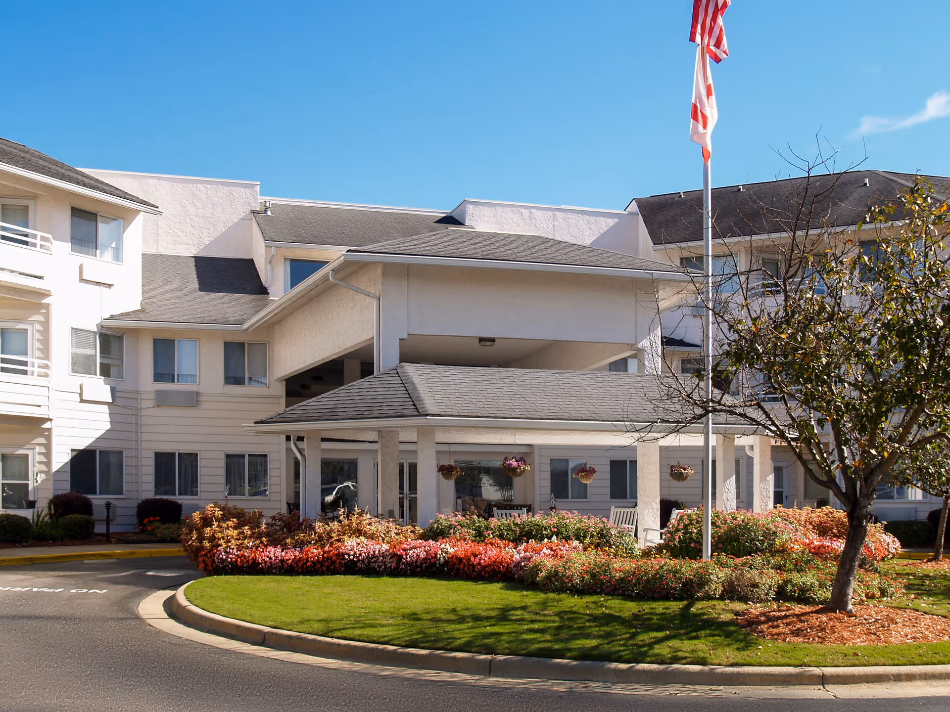 Exterior view of Asher Point Independent Living of Montgomery showing the entrance with a covered drop-off area, surrounded by well-maintained landscaping including colorful flowers, green grass, and a tree. An American flag is flying on a flagpole near the entrance under a clear blue sky.