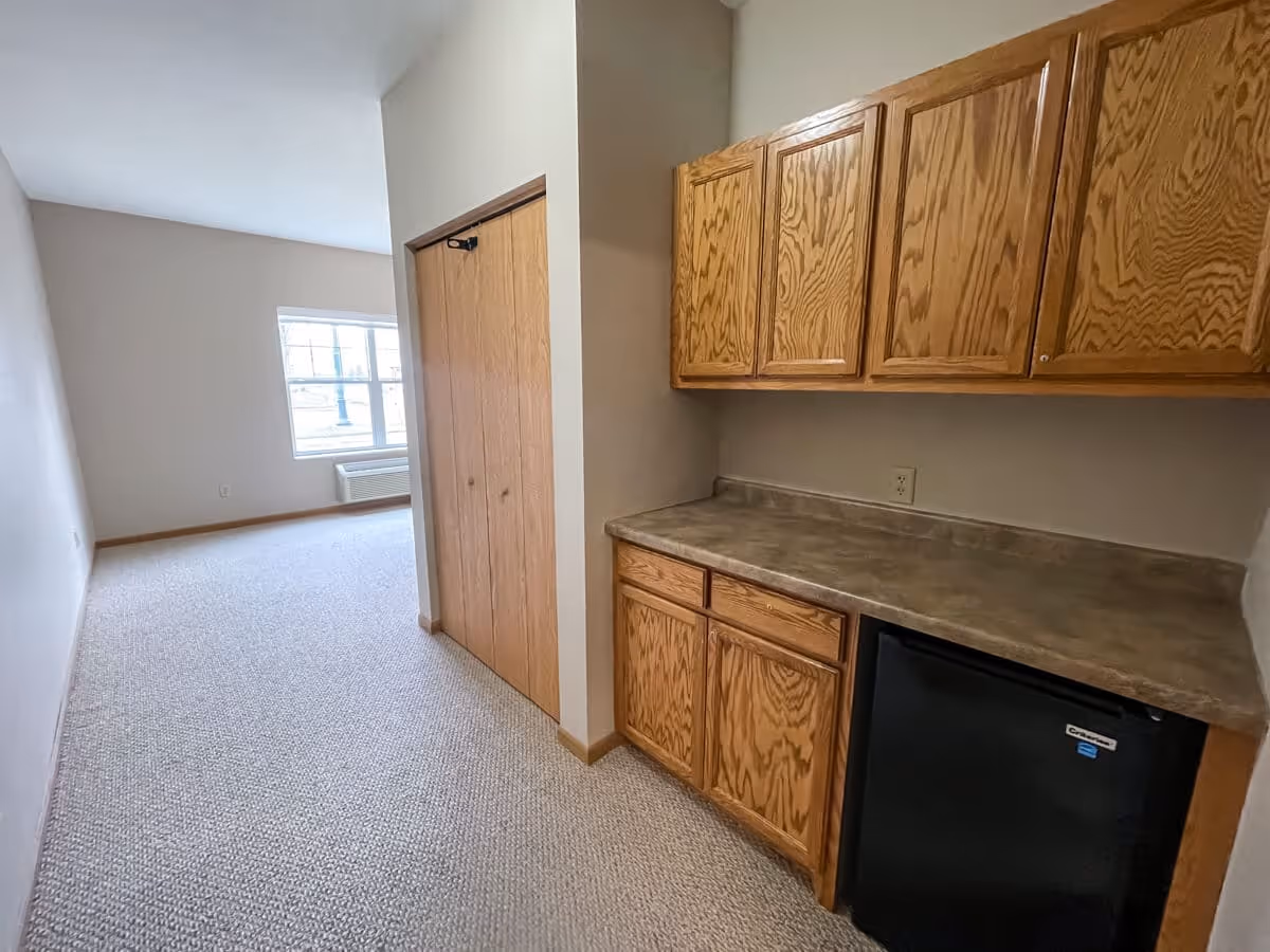 Interior view of a room in Lino Lakes Assisted Living featuring a small kitchenette area with wooden cabinets, a countertop, and a mini refrigerator. Adjacent to the kitchenette is a hallway leading to a window with natural light and a closet with wooden double doors. The room has beige walls and carpeted flooring.