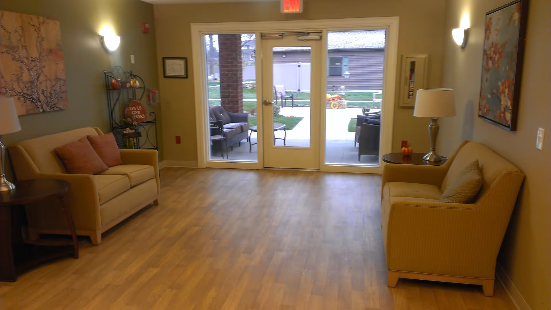 A cozy seating area inside a senior care facility with two beige sofas facing each other, each with cushions. There are side tables with lamps on either side of the sofas. The room has wooden flooring and light-colored walls adorned with framed artwork. At the far end, glass double doors lead to an outdoor patio area with additional seating and greenery visible outside.