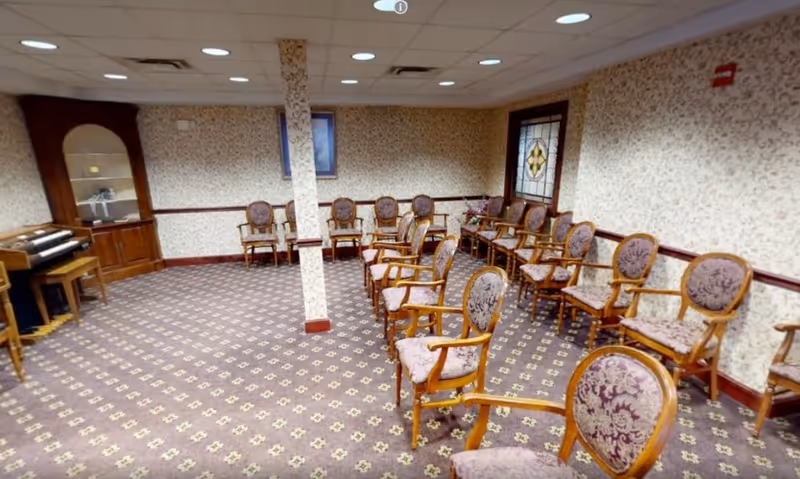 Carpeted community/activity room with rows of upholstered wooden chairs around the perimeter, patterned wallpaper, a small piano and a stained-glass window.