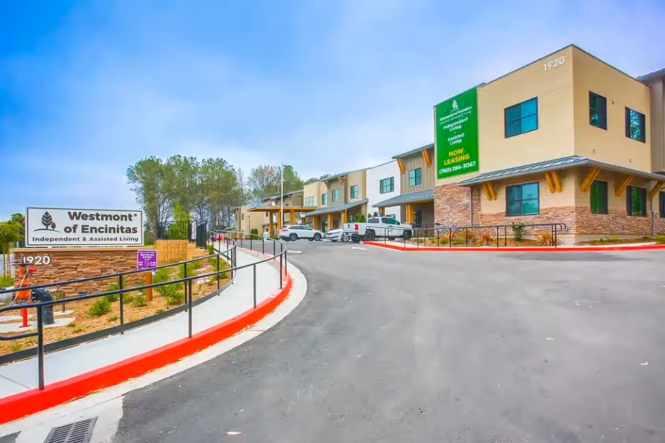 Exterior view of Westmont of Encinitas, an independent and assisted living facility, showing the building, parking area with vehicles, and signage with the facility name and leasing information.