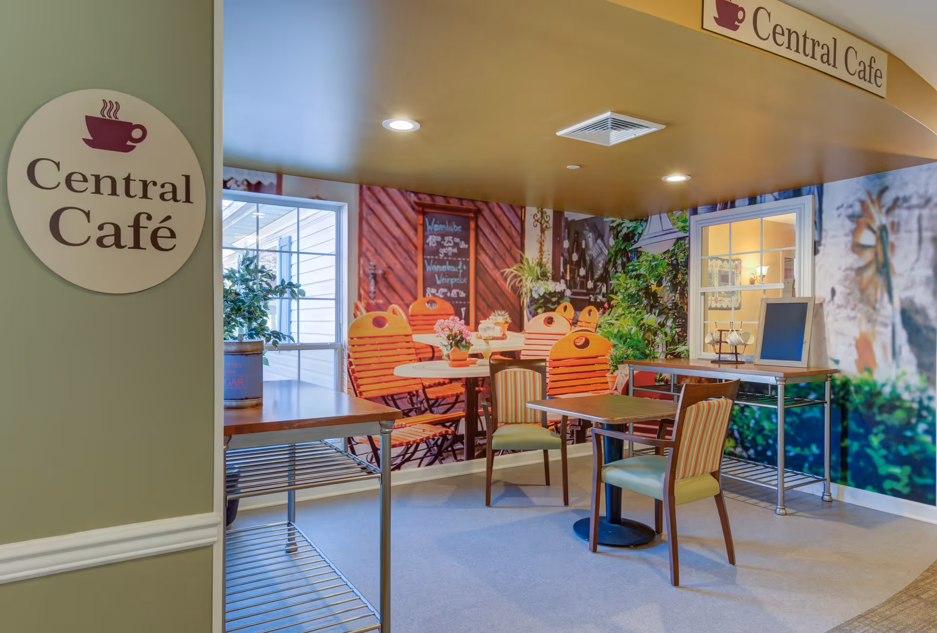 Interior view of a small café area named Central Café with a sign on the wall. The space includes a table with two chairs, a counter with a small chalkboard, and a large wall mural depicting an outdoor café scene with orange chairs, tables, plants, and a chalkboard menu.