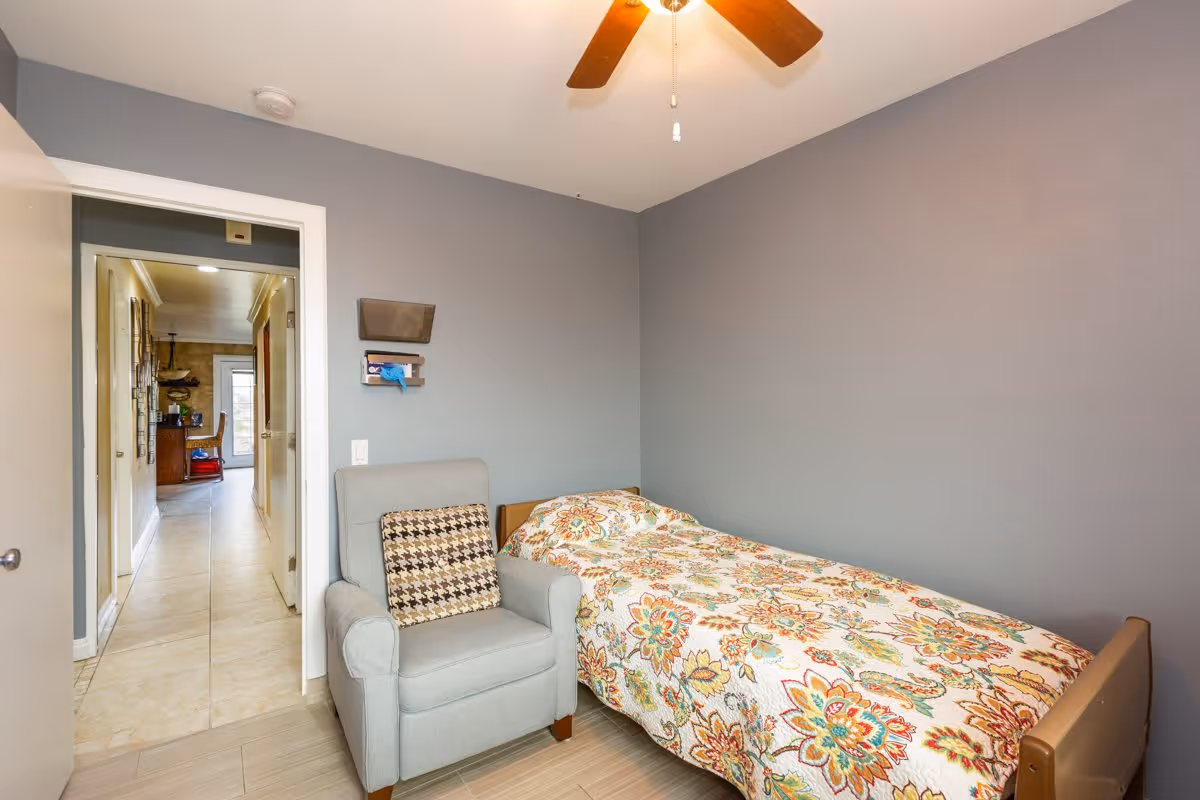 A small bedroom with a single bed covered in a colorful floral quilt and a gray armchair with a patterned cushion. The walls are painted gray, and there is a ceiling fan with wooden blades above. The door is open, showing a hallway leading to another room with tiled floors.