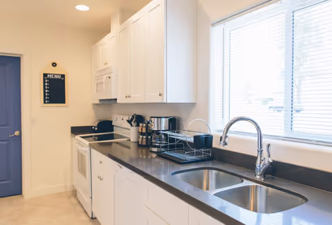 A clean and modern kitchen with white cabinets, a black countertop, a double sink with a chrome faucet, a dishwasher, a white stove with an oven, a microwave, and a window with blinds letting in natural light. A small blackboard menu hangs on the wall near a blue door.