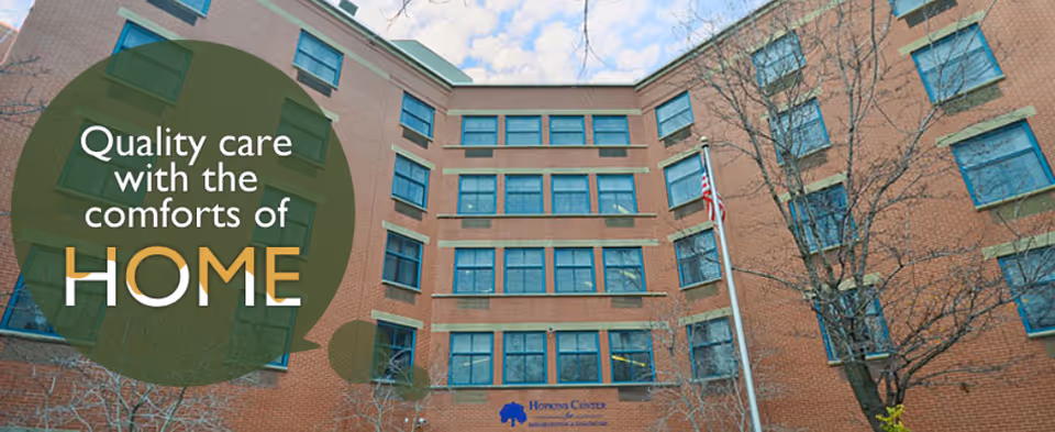Exterior view of a multi-story brick building with many windows, a flagpole with an American flag, and leafless trees in front. Text overlay reads 'Quality care with the comforts of HOME'.