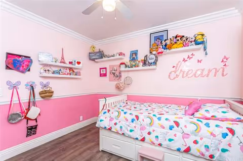 Pink-themed bedroom with a single daybed, colorful patterned bedding, wall shelves holding toys and decor, and a 'dream' wall decal.