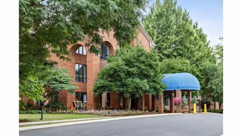 Exterior view of a brick building with arched windows partially obscured by green trees and shrubs. There is a blue awning over the entrance, with flower pots hanging nearby. The scene includes a paved driveway and well-maintained landscaping.