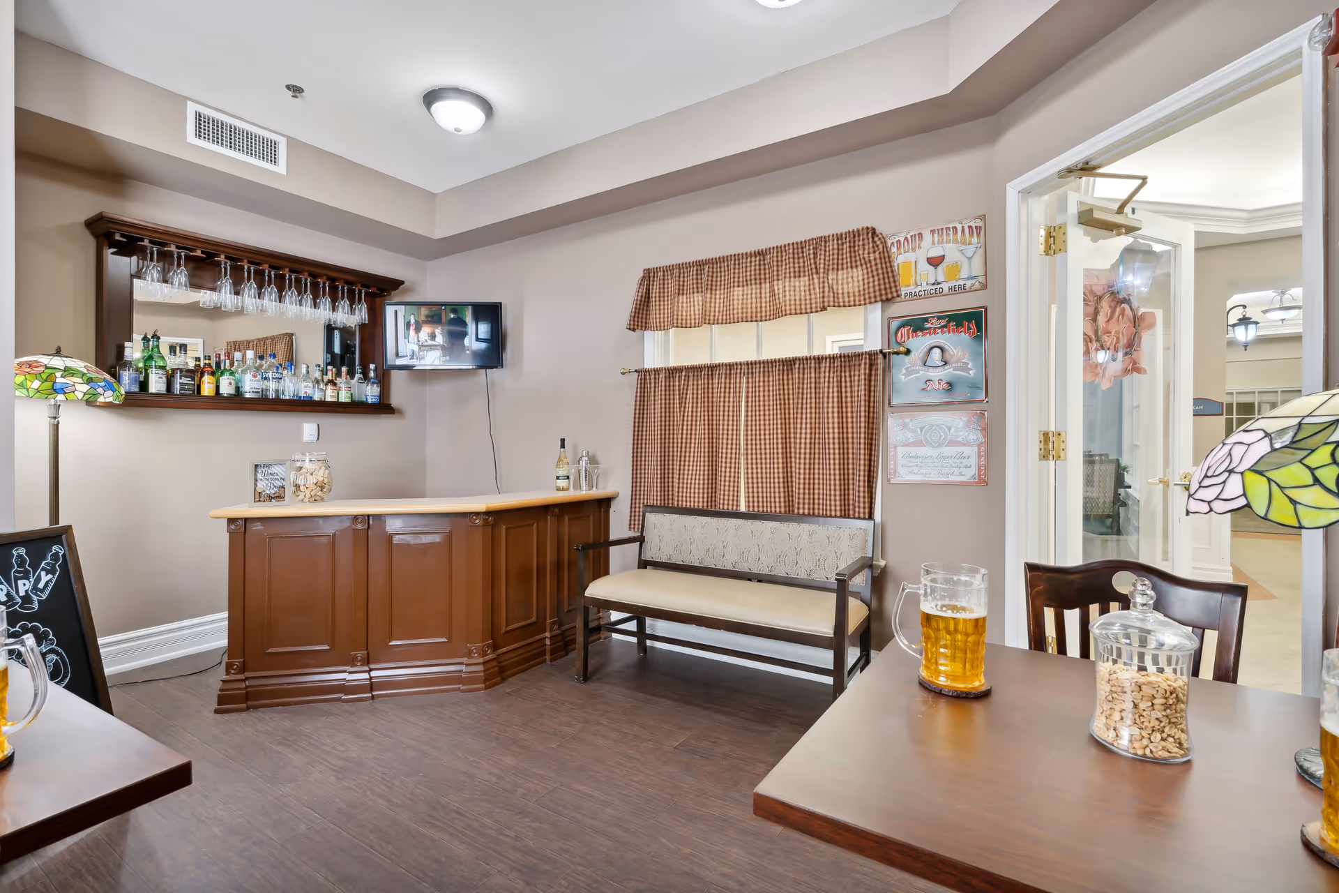 Interior view of a cozy assisted living facility lounge area featuring a wooden bar with various bottles and hanging wine glasses, a small wall-mounted TV, a cushioned bench with checkered curtains behind it, and a table with a pitcher of beer and a jar of snacks. The room has warm lighting and decorative lamps with stained glass shades.