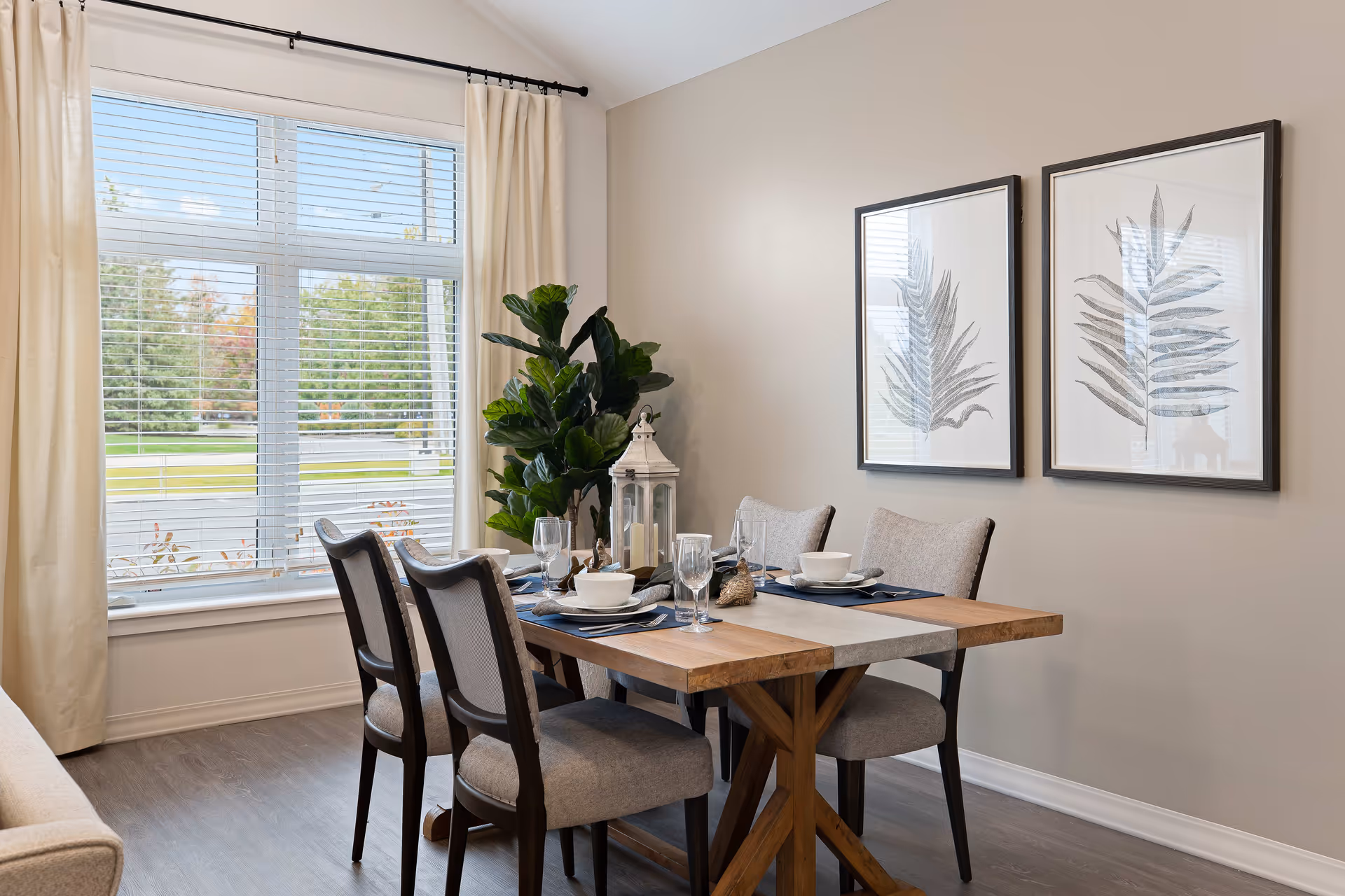 A dining area with a wooden table set for four, featuring gray cushioned chairs, white bowls, glasses, and silverware. The room has a large window with white blinds and cream curtains, a green potted plant in the corner, and two framed botanical prints on the beige wall.