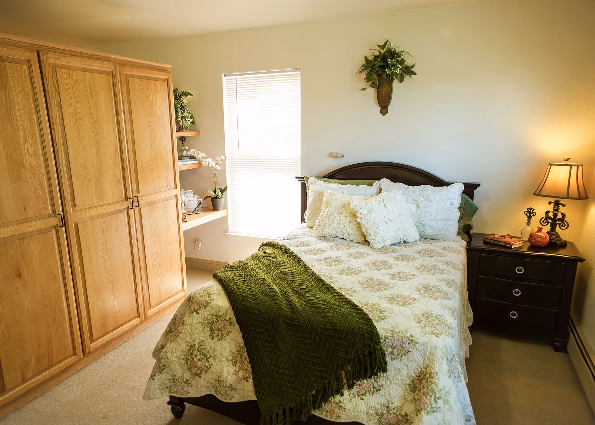 A cozy bedroom with a bed covered in floral bedding and multiple pillows. A green throw blanket is draped across the foot of the bed. To the right of the bed is a dark wooden nightstand with a lamp, books, and a small decorative pumpkin. On the left side of the room, there is a large wooden wardrobe and a small corner shelf with plants and decorative items. A window with blinds is centered on the back wall, and a wall-mounted plant adds greenery above the bed.