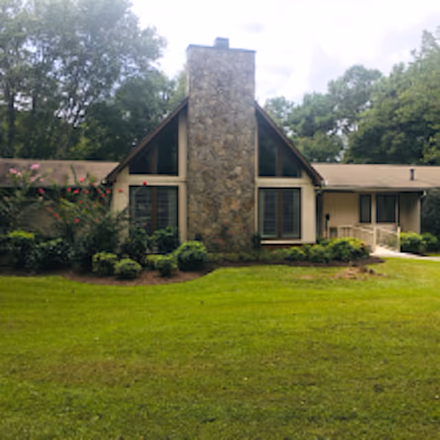 Front view of a single-story house with a central stone chimney, lawn and surrounding trees.